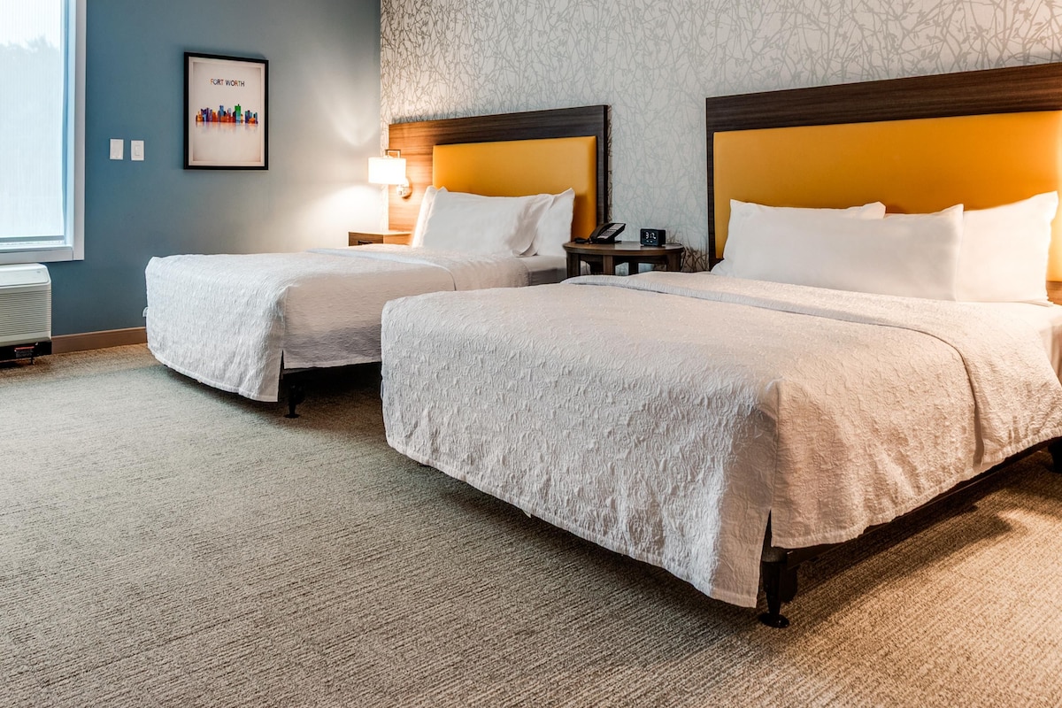 Habitación de hotel con dos camas, una mesita de noche con lámpara, un cuadro enmarcado en la pared y una ventana.