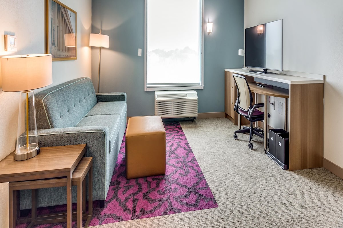 Une chambre d’hôtel avec un canapé, un bureau avec un ordinateur, une télévision, une fenêtre donnant sur un paysage agréable et une lampe.