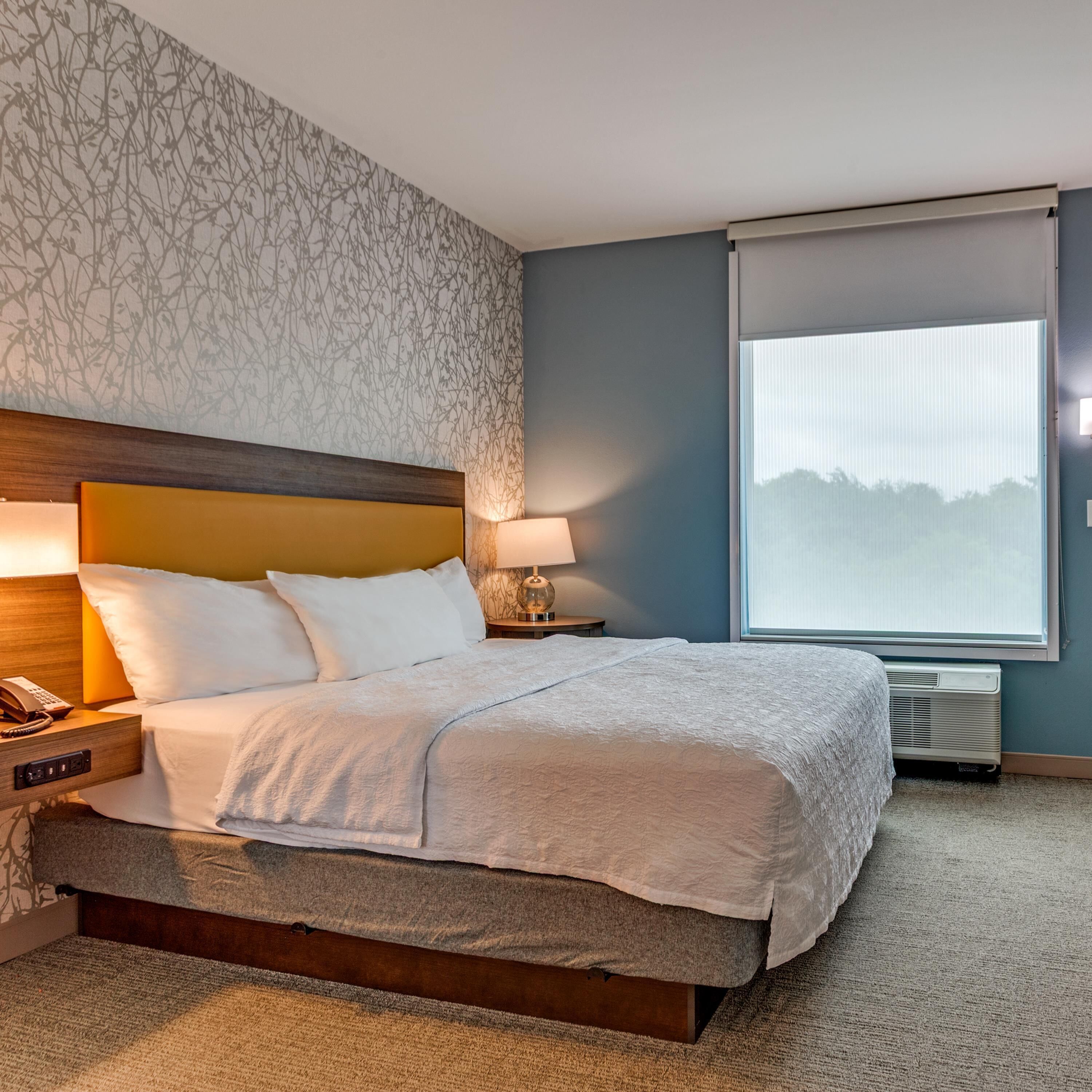 A neatly made bed with a headboard, a bedside lamp, a phone on the nightstand, and a large window with blinds.