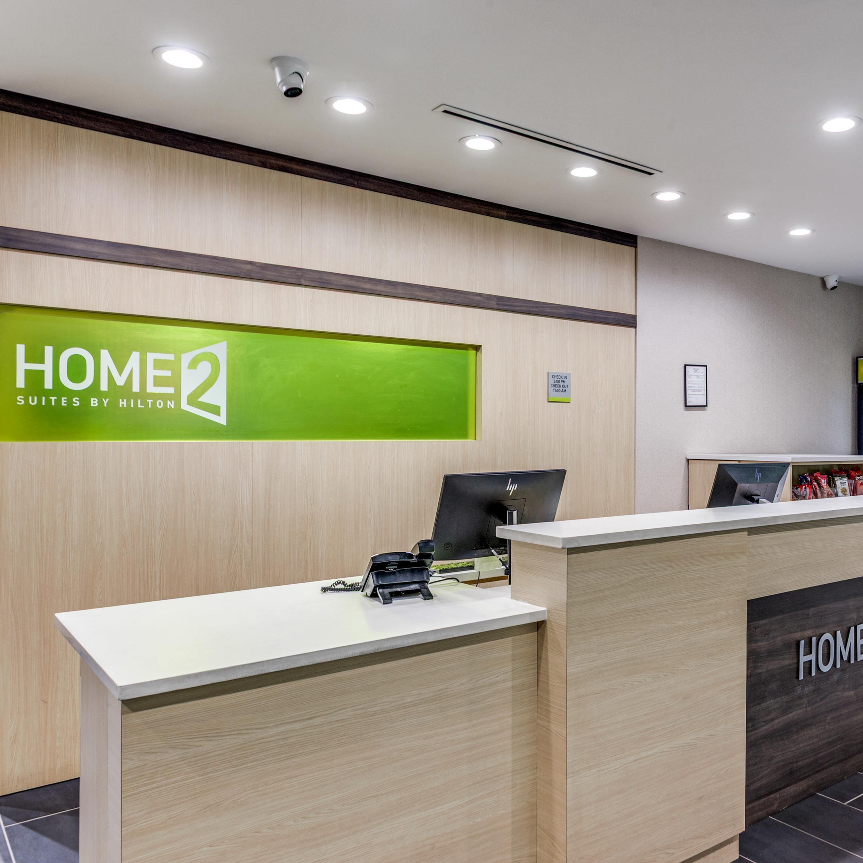 A hotel reception desk with a green sign reading 'HOME2 SUITES BY HILTON,' a computer monitor, and a telephone.
