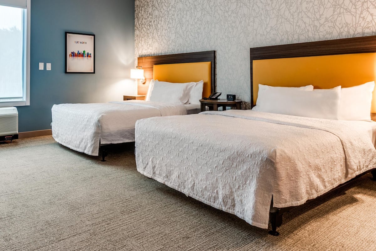 Une chambre d’hôtel avec deux lits, une table de chevet avec une lampe, un tableau encadré au mur et une fenêtre.