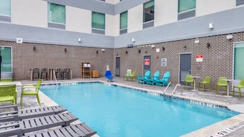 Una piscina interior con sillas y mesas de descanso, rodeada por un muro de ladrillo con ventanas y señales de salida de emergencia.