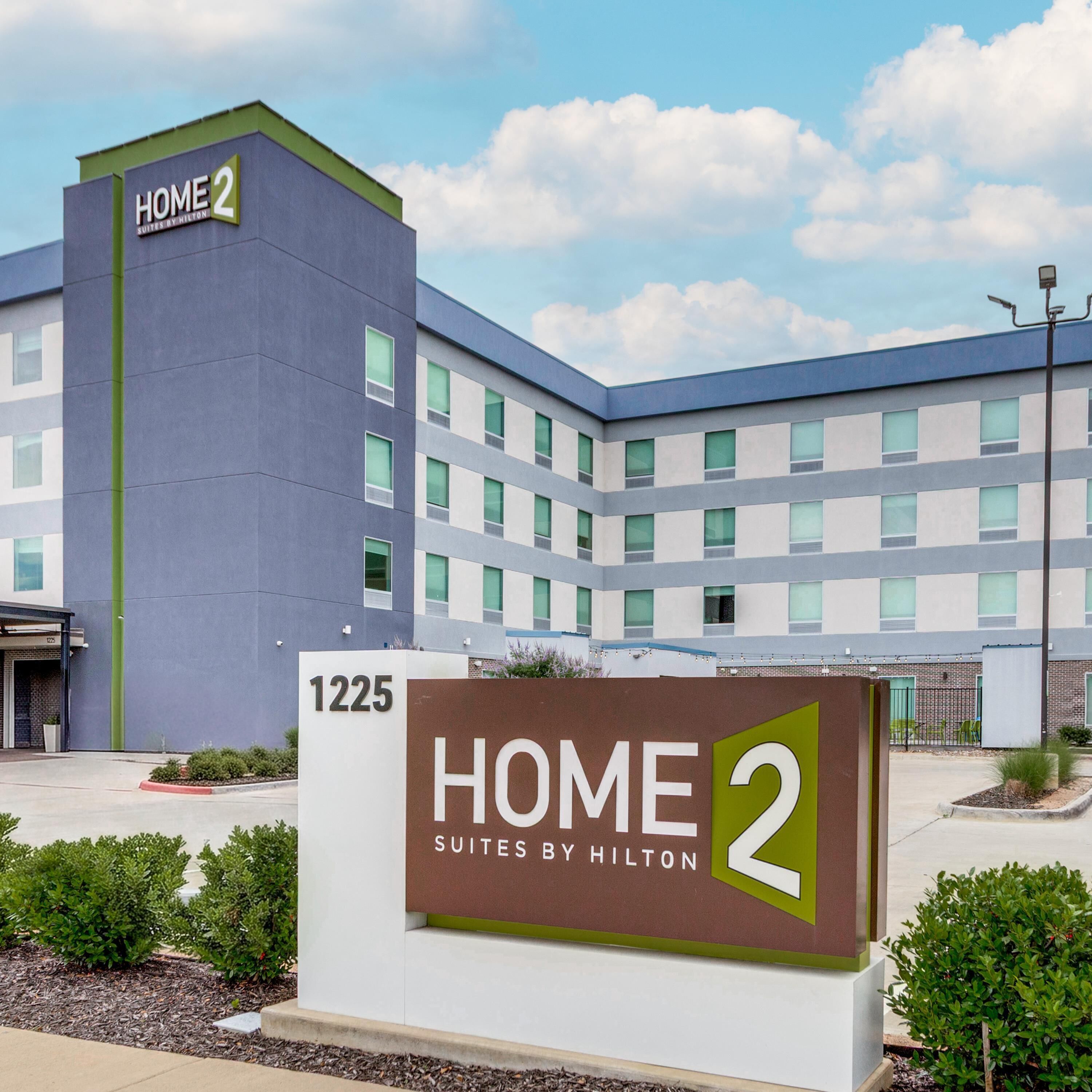 A modern hotel building with a sign reading 'HOME 2 SUITES BY HILTON' and the address '1225'.