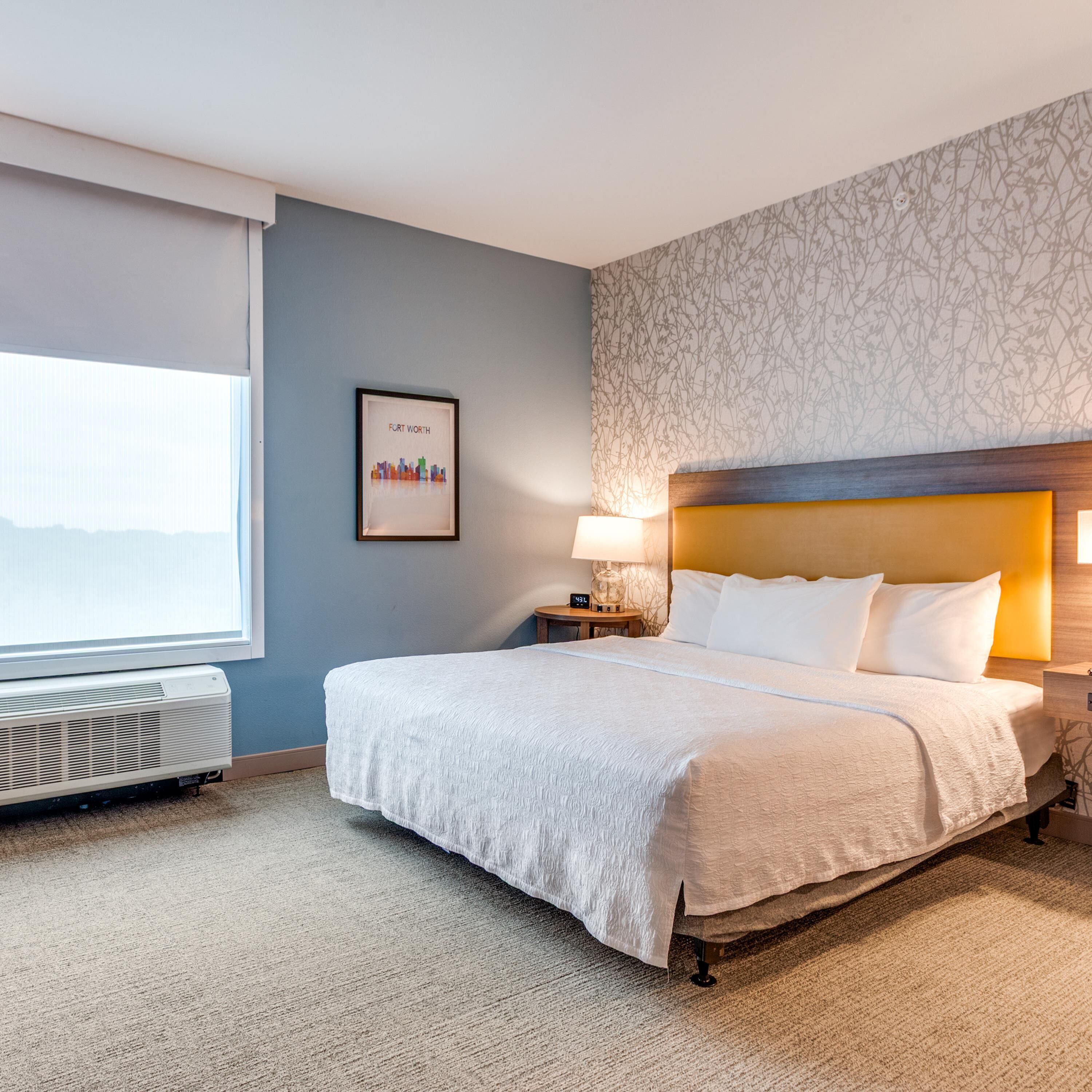 A hotel room with a bed, a nightstand with a lamp, a framed picture on the wall, and a window with blinds.