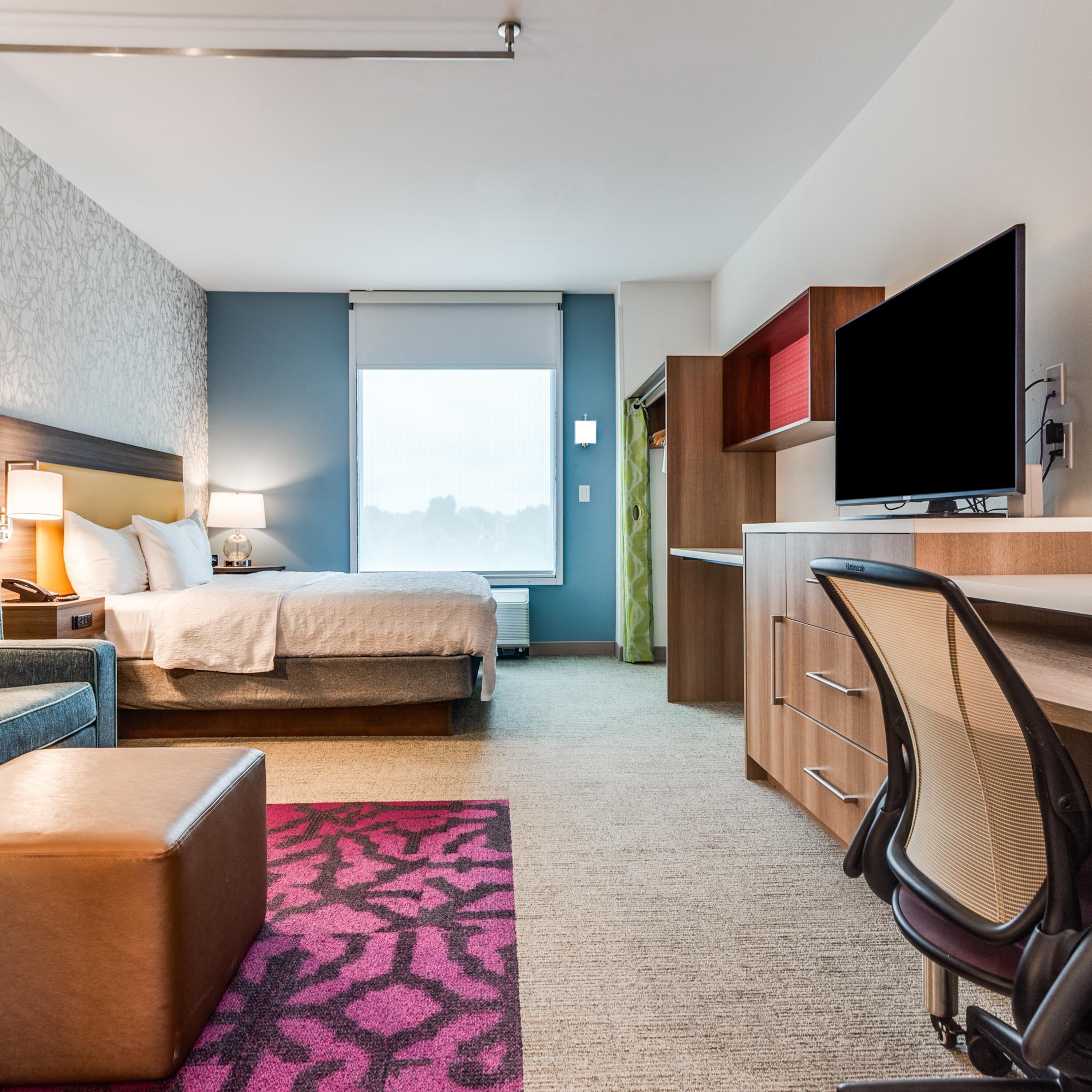 Habitación de hotel con cama, escritorio, silla, televisión y un ventanal grande.