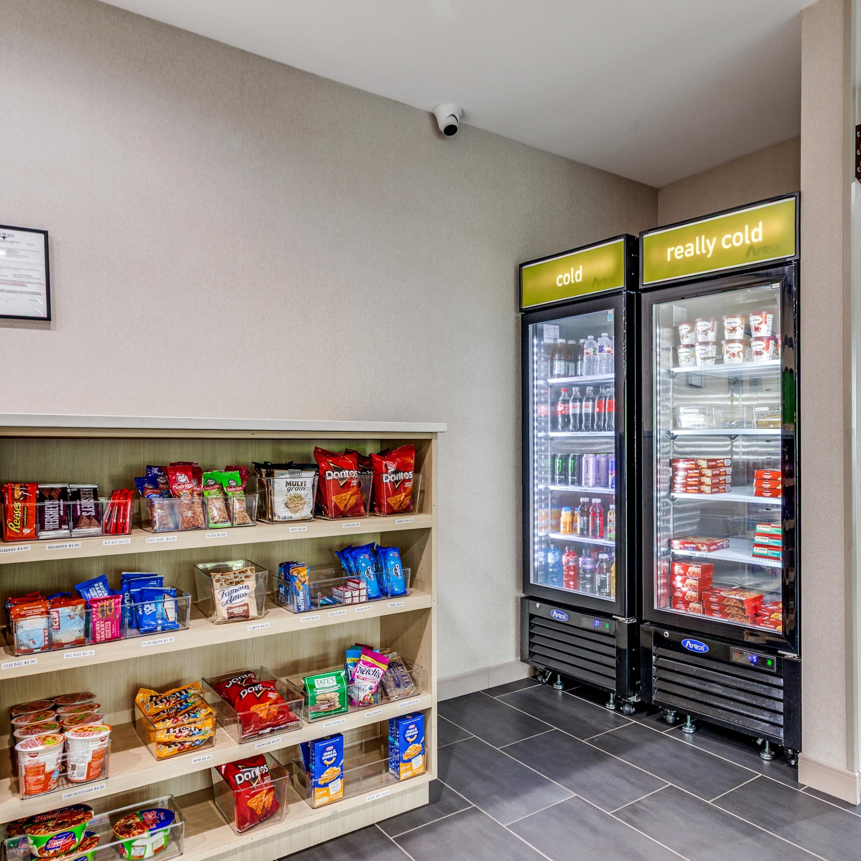 Zona de venta automática de aperitivos y bebidas con varias estanterías llenas de diferentes opciones y un refrigerador de dos puertas con un letrero que indica 'muy frío'.