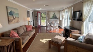 Amplia sala de estar con sofá, sillones, una mesa de centro y televisor. Hay una zona de comedor con mesa y sillas, y un ventanal con cortinas.
