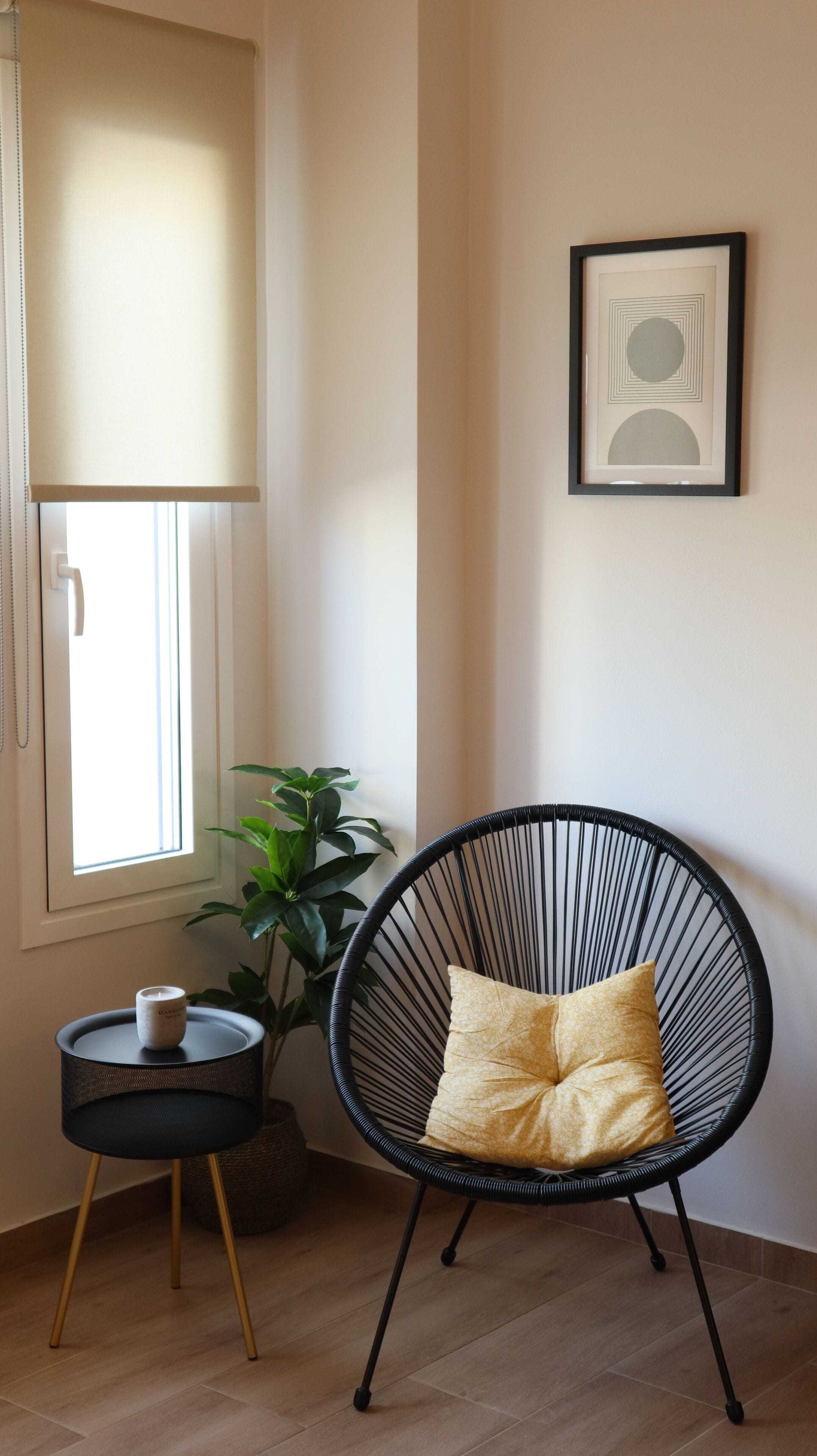 A room with a black wicker chair, a small round table with a candle, a potted plant, and a framed artwork on the wall.