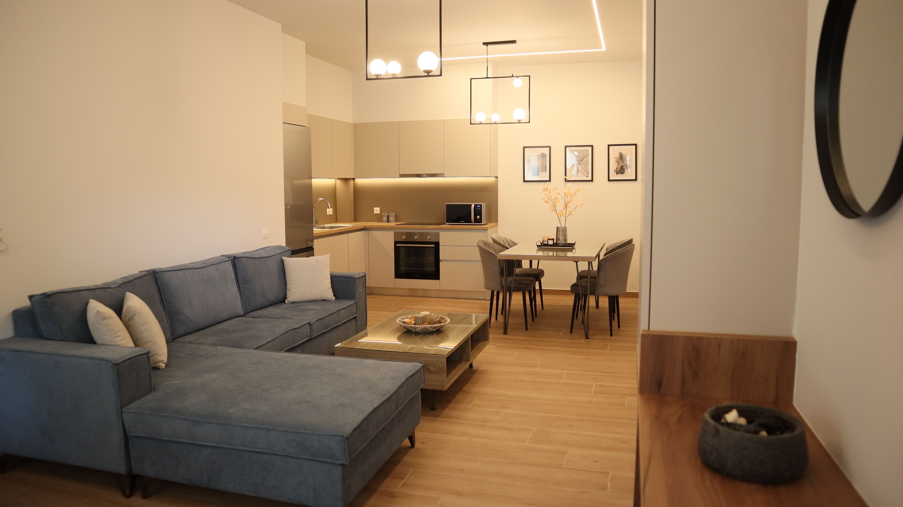 A modern living room with a grey sectional sofa, a dining area with a wooden table and chairs, and a kitchen with a microwave and oven.