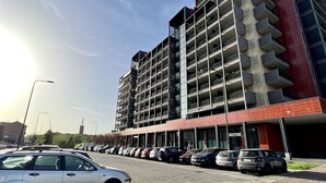Ein mehrgeschossiges Gebäude mit roter und grauer Fassade, ein Parkplatz mit mehreren Autos und einem Pflastergrund.