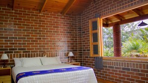 Una habitación con pared de ladrillo, techo de madera, un ventanal grande, una cama con colcha blanca y dos mesitas de noche con lámparas.