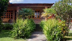 Una casa de ladrillo con tejado de baldosas, un camino de piedra y abundante vegetación, incluyendo helechos y plantas con flores.