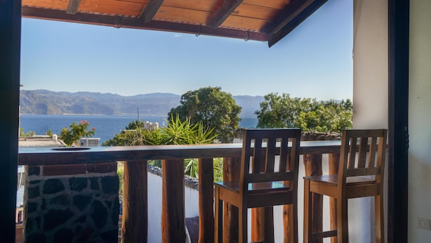 Un balcón con sillas y mesa de madera, con vistas panorámicas a montañas y una masa de agua.