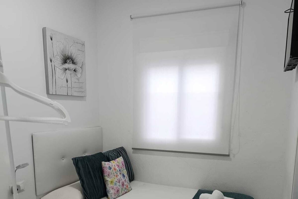 A single-bed room with a white headboard, a window with blinds, a bed with a green throw blanket, a pillow with a floral pattern, and a small bedside table with a remote control.