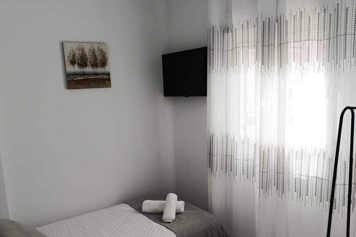 A minimalist bedroom with a bed, a flat-screen TV, a painting, and sheer curtains with hanging elements.