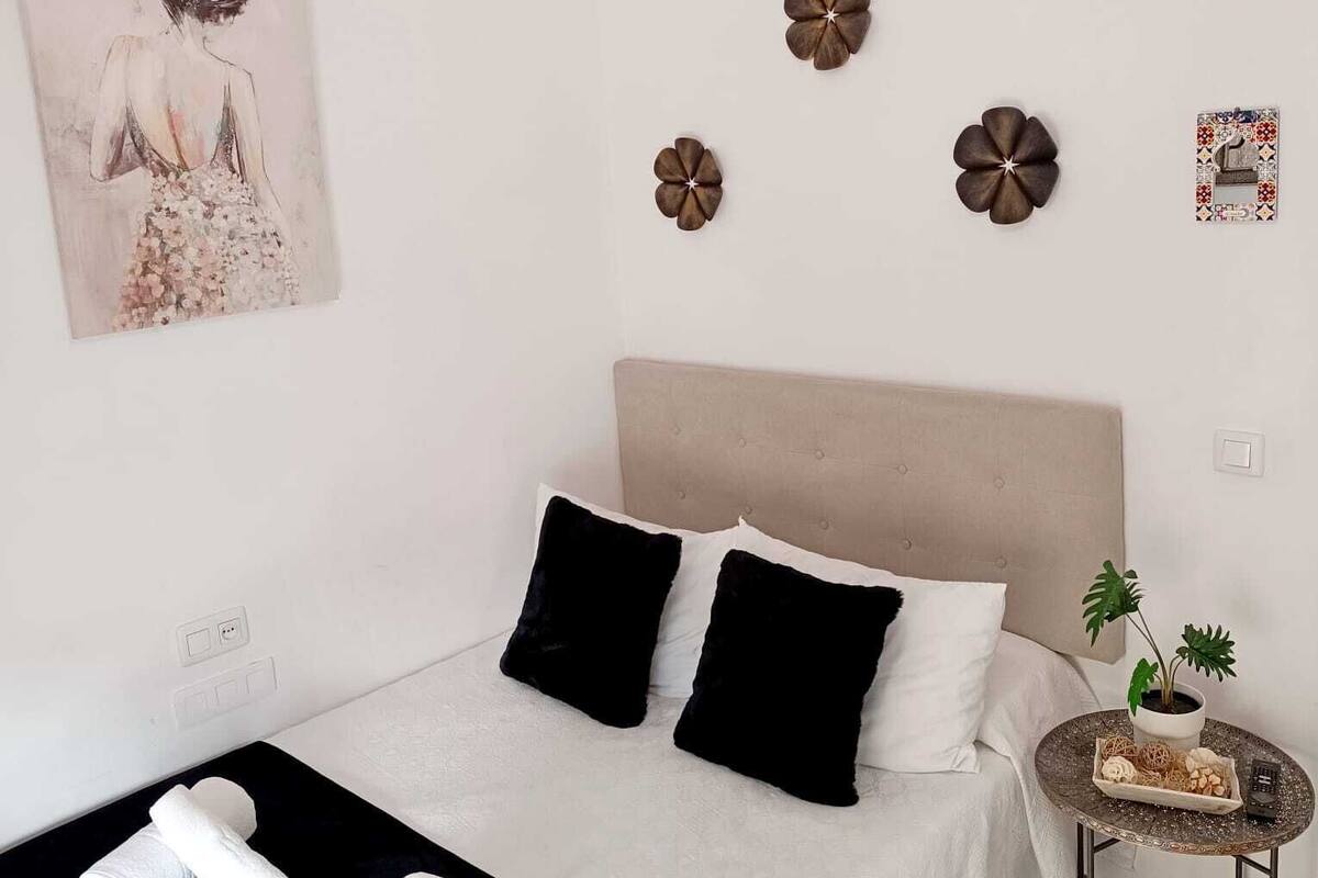 A bedroom with a bed, pillows, a bedside table with a plant, and a wall-mounted light fixture.