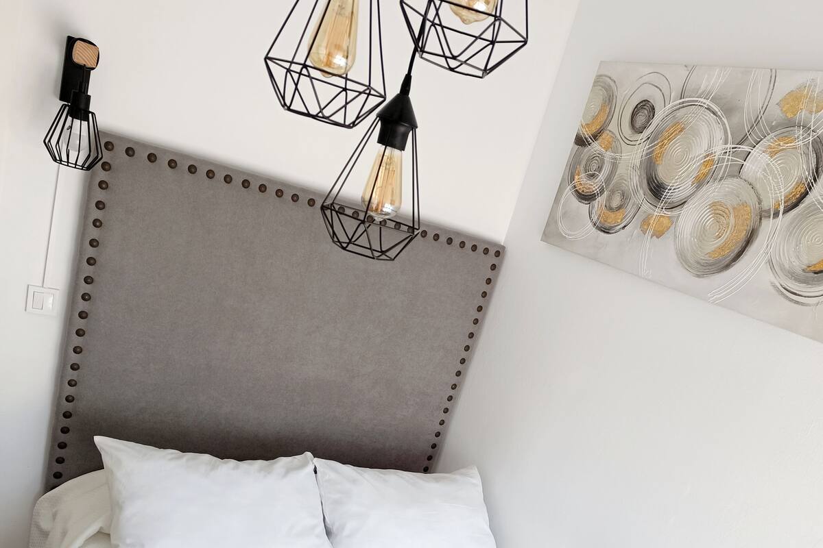 A neatly made bed with white linens and a patterned throw, a headboard with a studded design, and a wall-mounted light fixture with exposed bulbs.