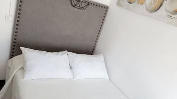 A neatly made bed with white linens and a patterned throw, a headboard with a studded design, and a wall-mounted light fixture with exposed bulbs.