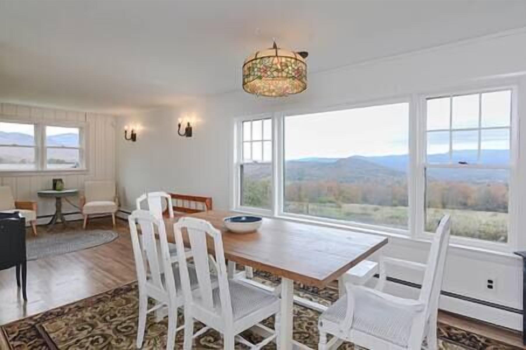 Un comedor con mesa de madera y sillas blancas, ventanas amplias con vistas al campo y una araña.