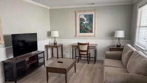 Ein Wohnzimmer mit Fernseher, zwei Nachttischen mit Lampen, einer Couch und einem gerahmten Bild an der Wand.