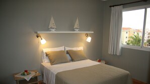 Un lit bien fait, recouvert d’une couette blanche et beige, avec deux lampes de chevet et une étagère sur laquelle se trouve une petite table portant une assiette de nourriture et une bouteille.