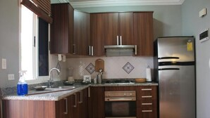 Une cuisine avec des meubles en bois, un réfrigérateur en acier inoxydable, un four encastré et une fenêtre avec des stores.
