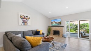 A modern living room with a grey sofa, yellow cushions, a wooden coffee table, a fireplace, and a TV mounted on the wall. - Lush Chic Living Private Backyard Heated Saltwater Pool (Indio)