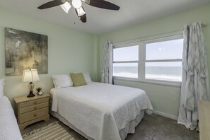Quarto com cama, mesa de cabeceira com abajur, janela com cortinas e vista para o oceano.
