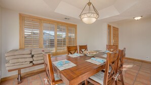 A dining area with a wooden table set for six, surrounded by chairs. There are window blinds on the left, a ceiling light fixture above, and a door on the right. - Well-appointed home just steps from the Colorado river! (Needles)