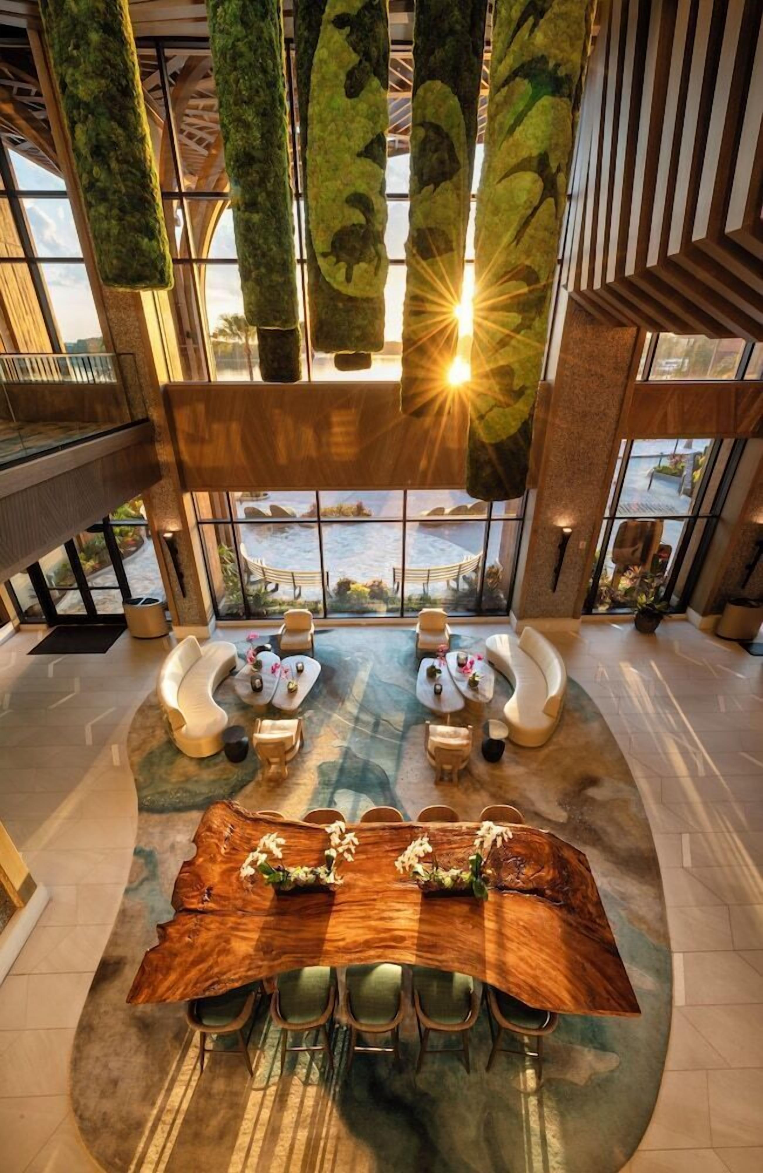 A spacious lobby with a large wooden table surrounded by chairs, a seating area with sofas, and a view of the outdoors through large windows.