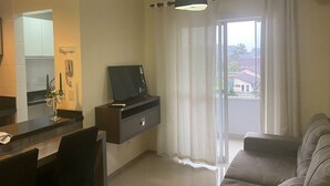 A living room with a sofa, a TV on a stand, a dining table with chairs, a window with curtains, and a view of buildings outside. - Loft Descolado Glória (Joinville)