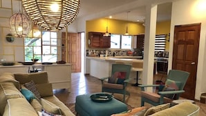 Ein geräumiges Wohnzimmer mit einer großen, beigefarbenen Couch, türkisfarbenen Sessel und einem Couchtisch. Im Hintergrund ist eine Küche mit Holzausstattung und einem großen Fenster zu sehen.