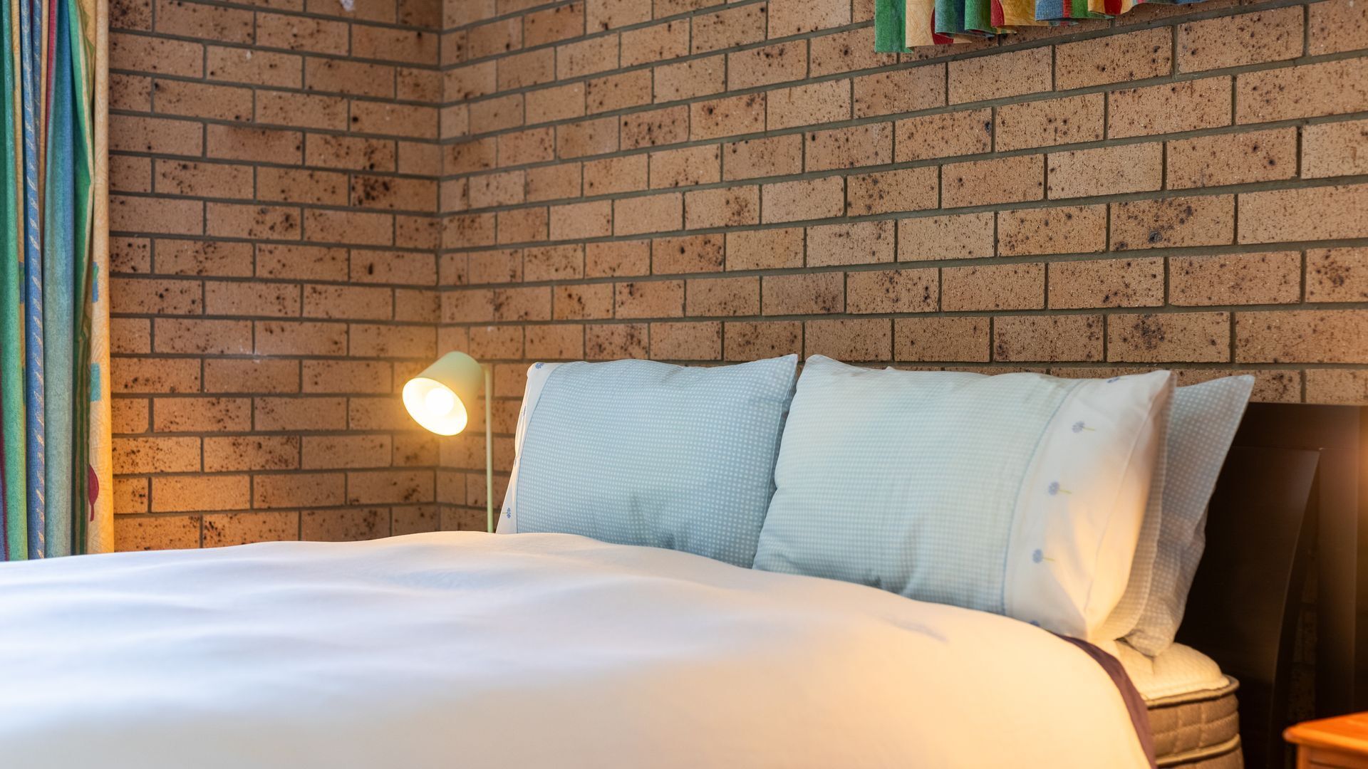 Una cama bien tendida con sábanas blancas en una habitación con pared de ladrillo y una lámpara de noche.