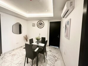 A dining area with a table and chairs, a wall clock, and an air conditioning unit. - Apex Suite (Accra)