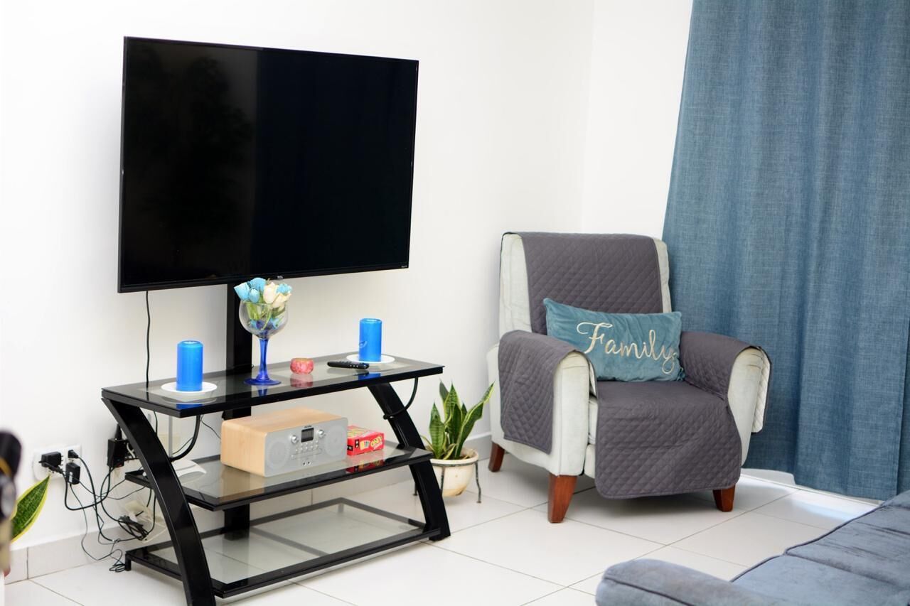 Ein Wohnzimmer mit einem an der Wand montierten Flachbildfernseher, einem Glastisch mit einer blauen Kerze, einer Topfpflanze und einem grauen Sessel mit einem Kissen mit der Aufschrift 'Family'.