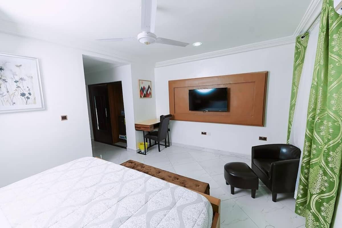 Ein Hotelzimmer mit einem Bett, einem Schreibtisch, einem an der Wand befestigten Fernseher, einem Deckenventilator und einem grünen Vorhang.