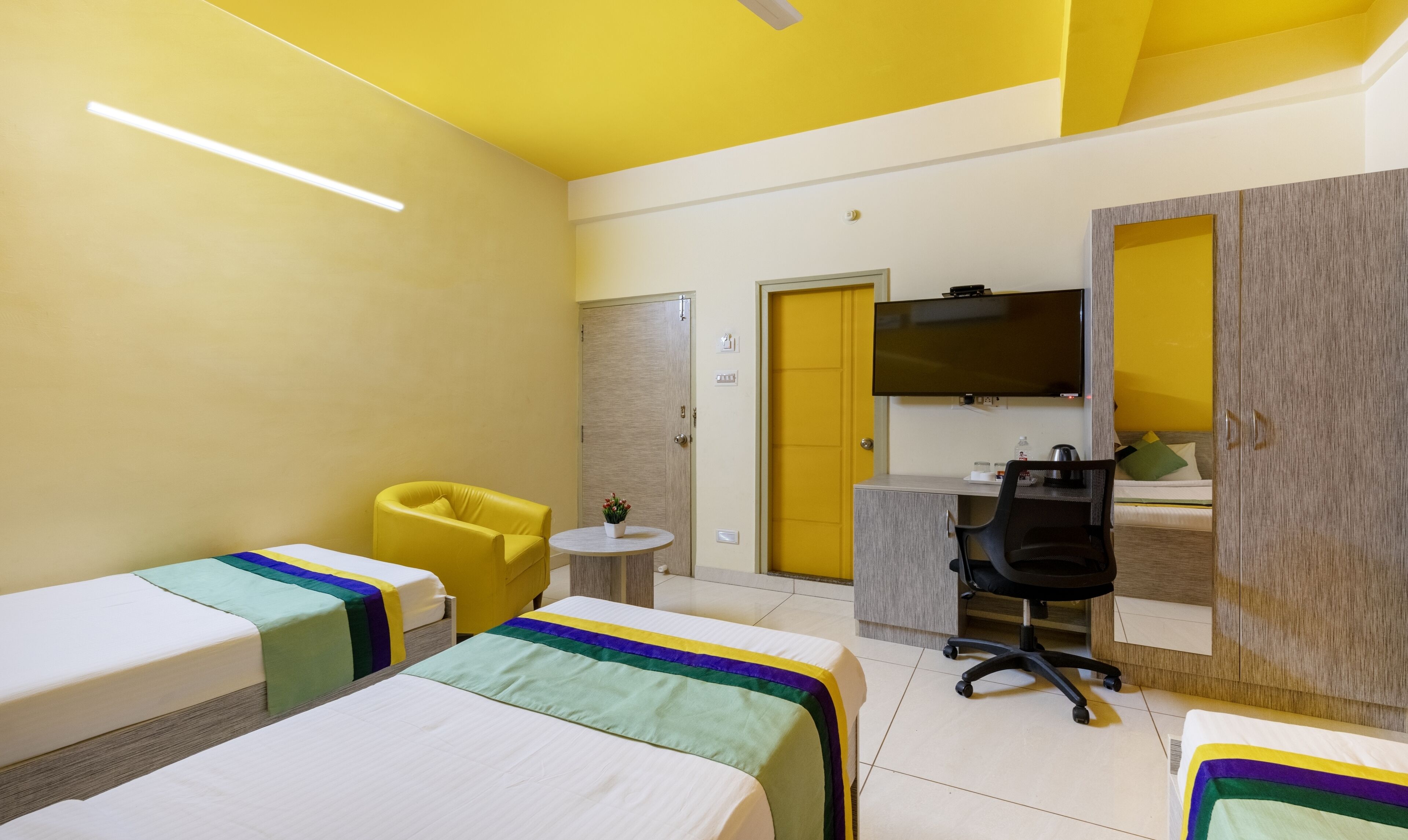 Een hotelkamer met twee enkele bedden, een klein rond tafeltje, een gele fauteuil, een bureau met stoel en een aan de muur bevestigde televisie.