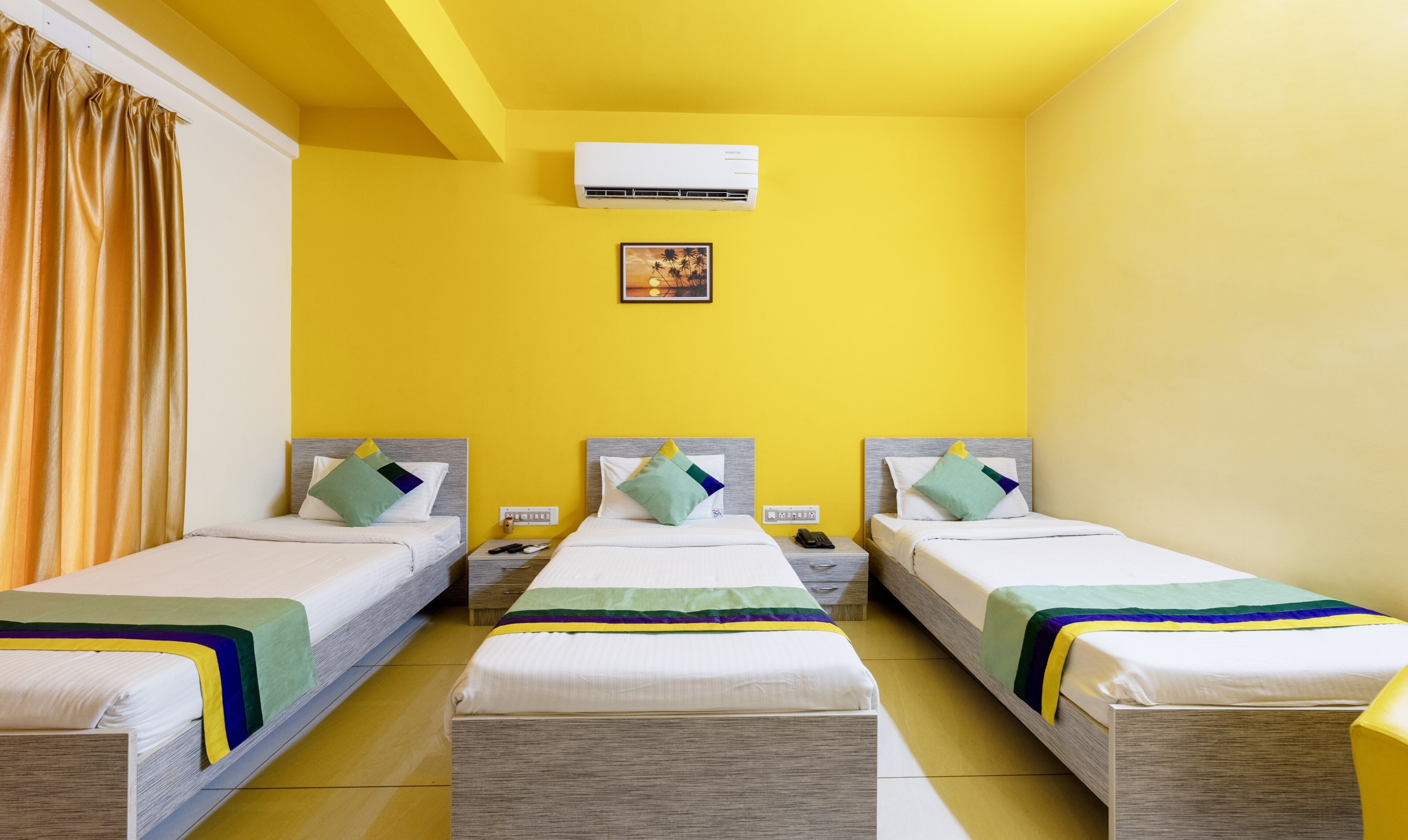 A hotel room with three single beds, each with a wooden headboard and a colorful bedspread. There is a wall-mounted air conditioner above the beds and a small framed picture on the wall.