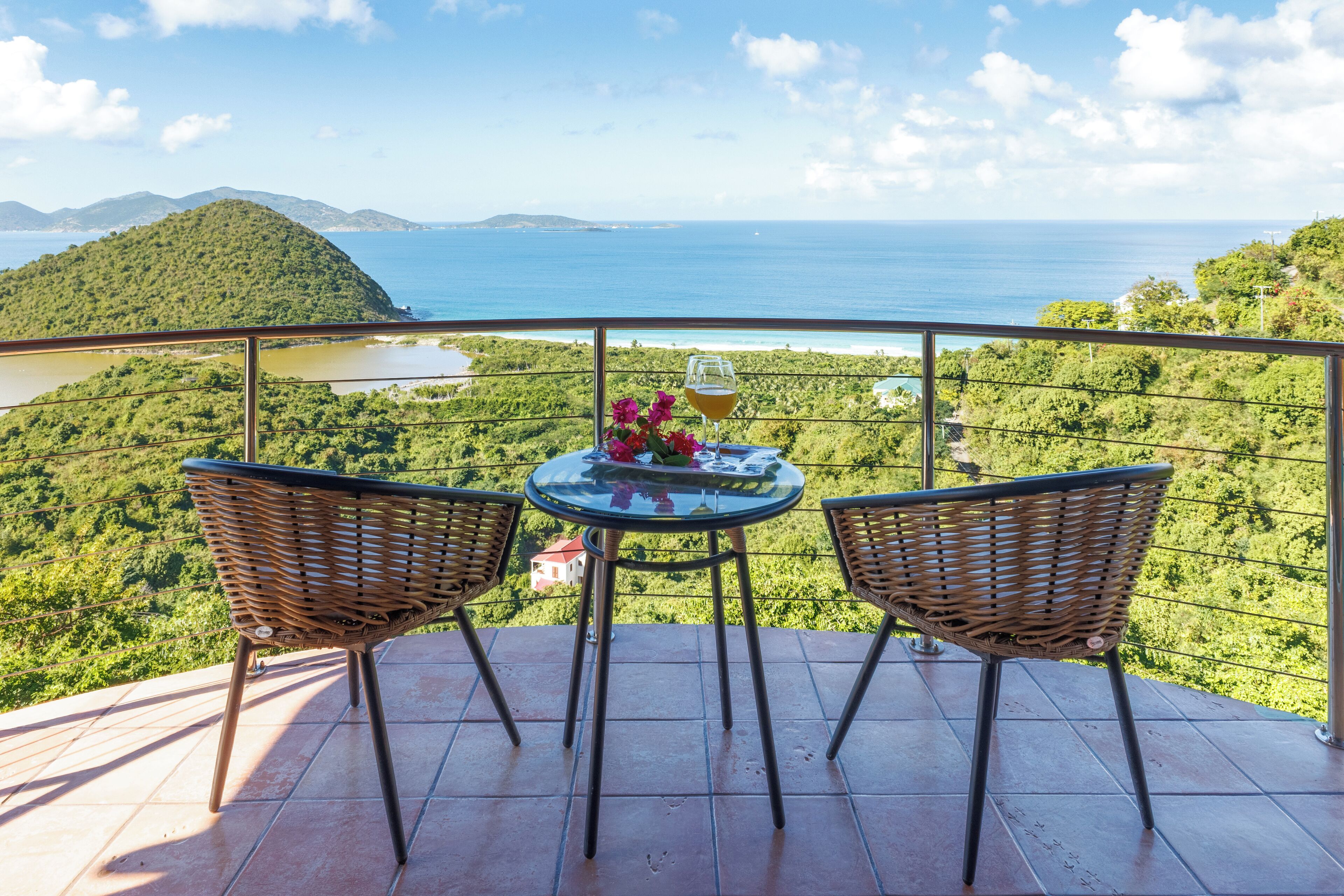 A balcony with wicker chairs and a table, overlooking a scenic landscape with hills and a body of water.