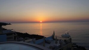 A sunset over a calm sea with a view of a coastal area, including buildings and a clear sky.