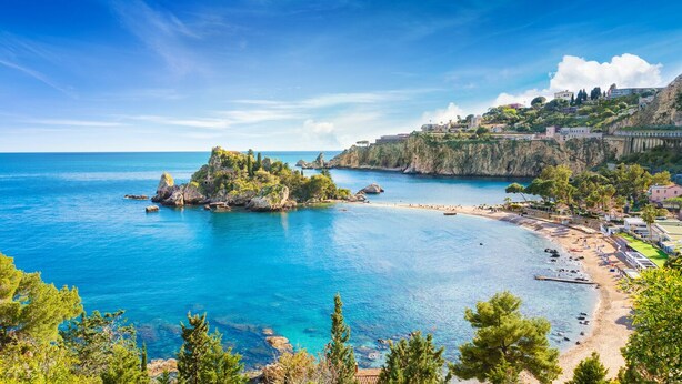 A coastal town with a clear blue sea, rocky outcrops, and a beach with buildings on a hillside.
