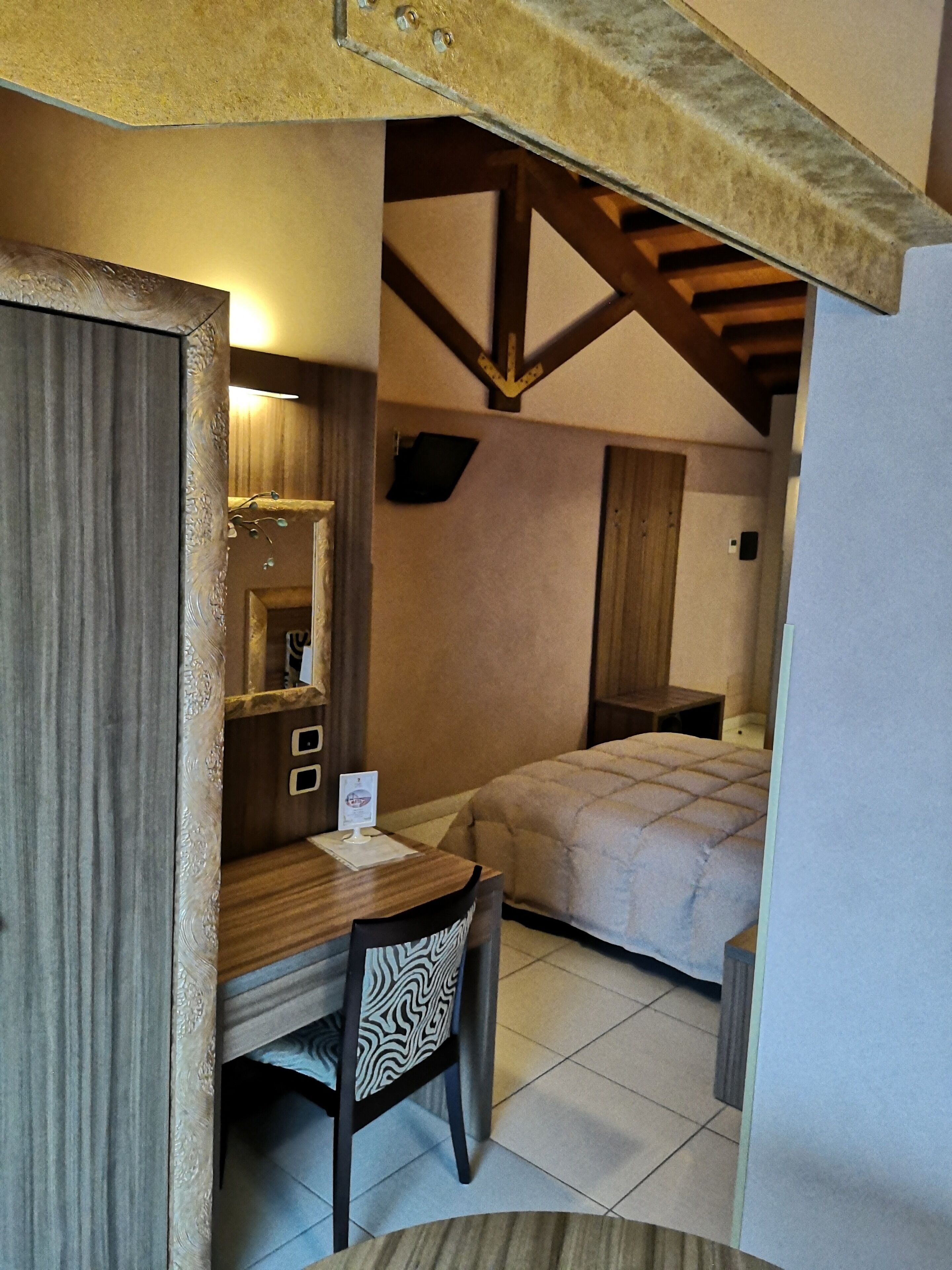 Una habitación compacta con cama, escritorio y silla. Hay una pequeña mesa con un cartel. La habitación tiene vigas de madera en el techo.