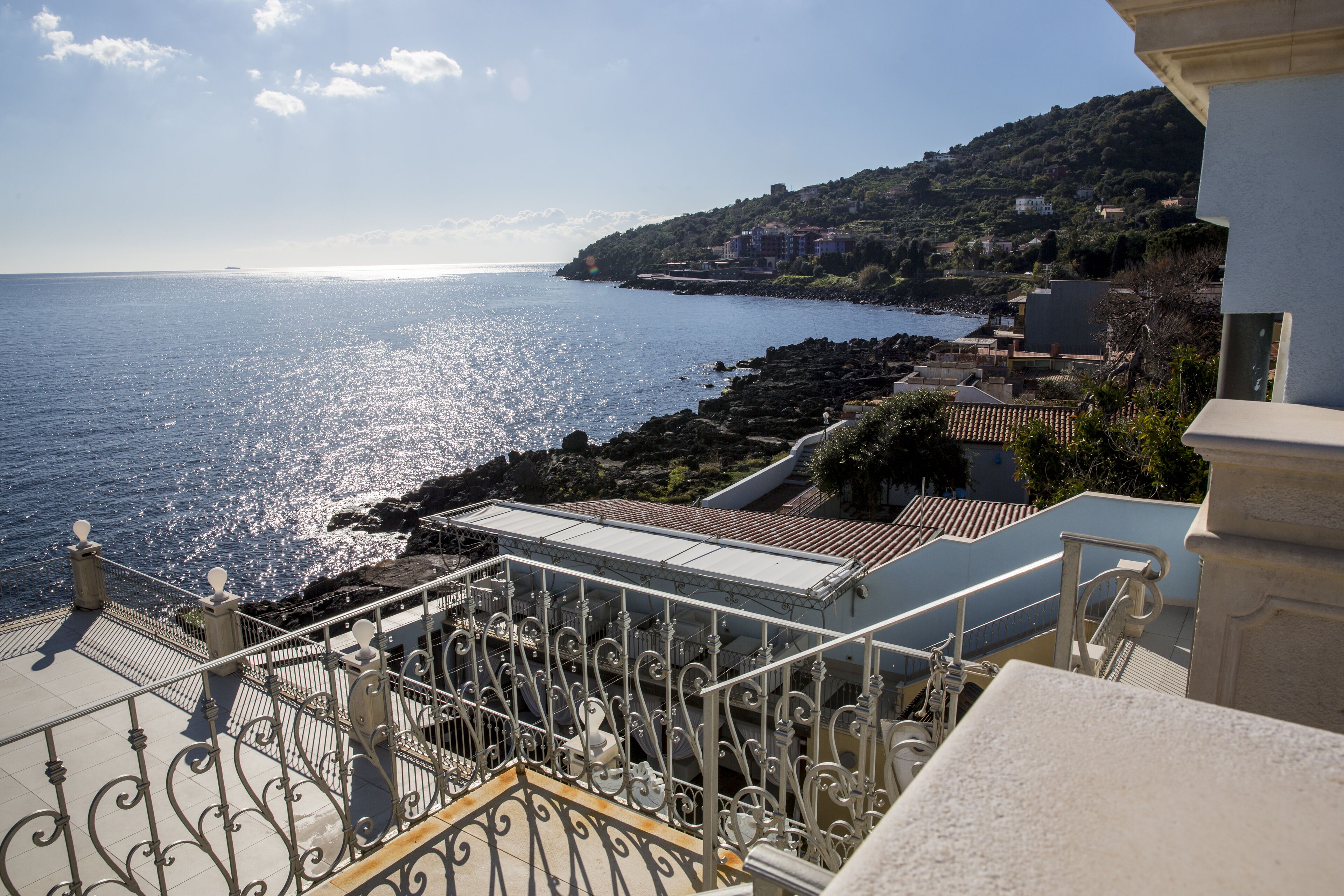 A coastal view from a balcony with a metal railing, a staircase, and a view of the sea and a hillside with buildings.