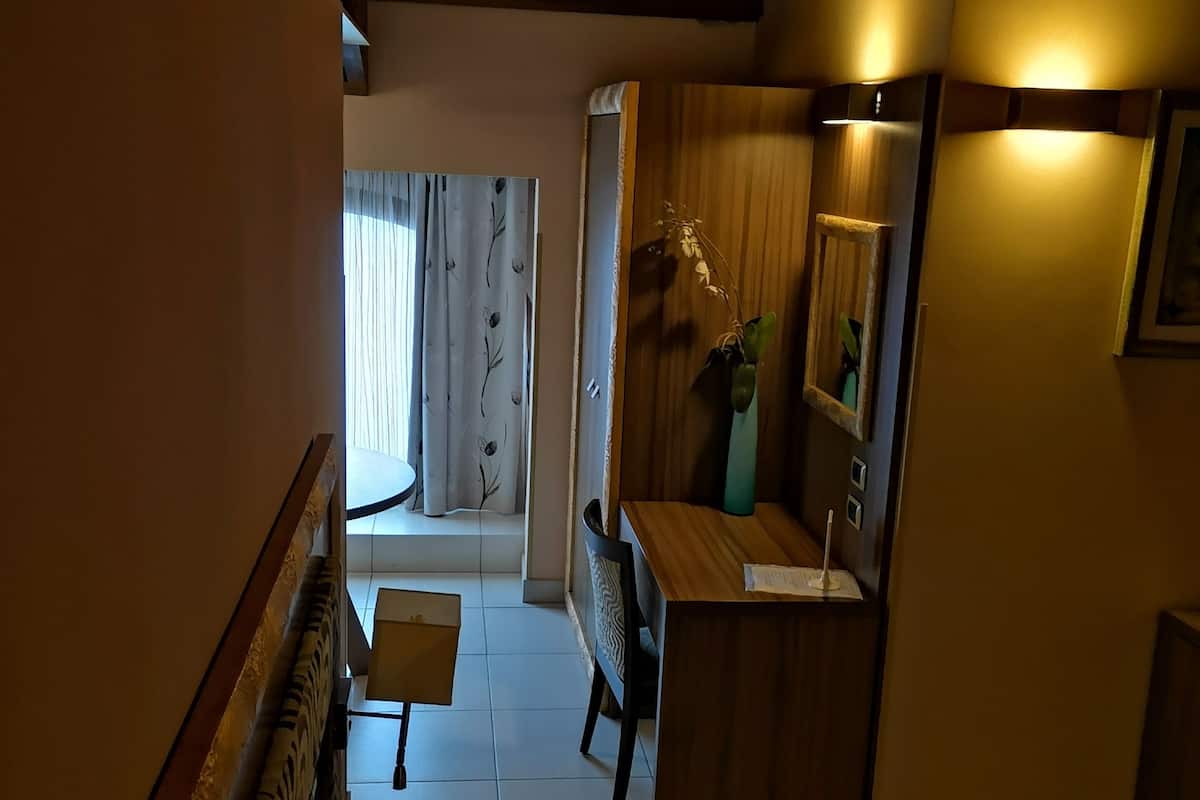 A modern living room with a sofa, a wooden coffee table, and a small table with a remote control. There is a wooden cabinet with a mirror and a vase with a plant. The room has a wooden ceiling and a window with curtains.