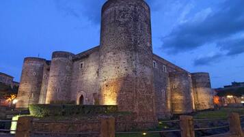 Un castillo de piedra histórico con múltiples torres y un gran torreón central, rodeado por un muro bajo y una zona cubierta de césped.