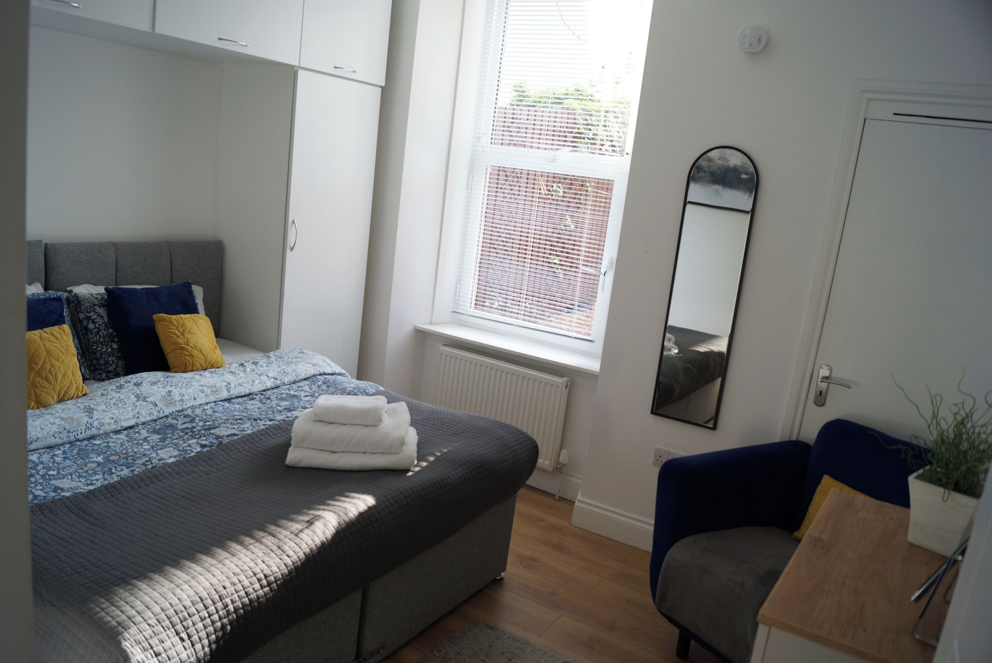 A bedroom with a bed, a chair, a mirror, and a window with blinds.