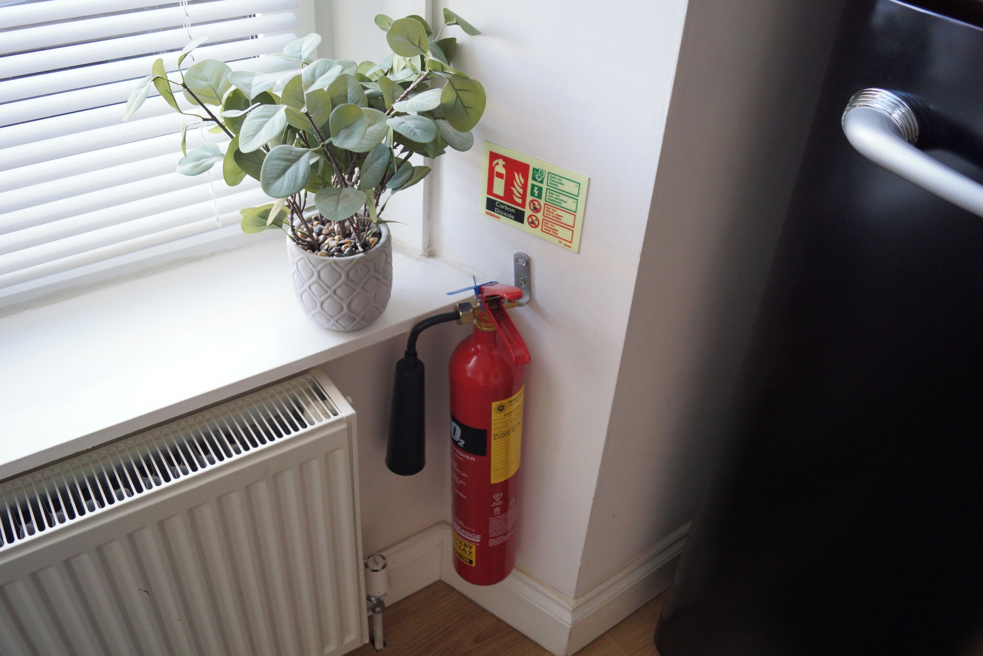 Ein Fenster mit Jalousien, ein Blumentopf, ein Feuerlöscher und eine Heizung.