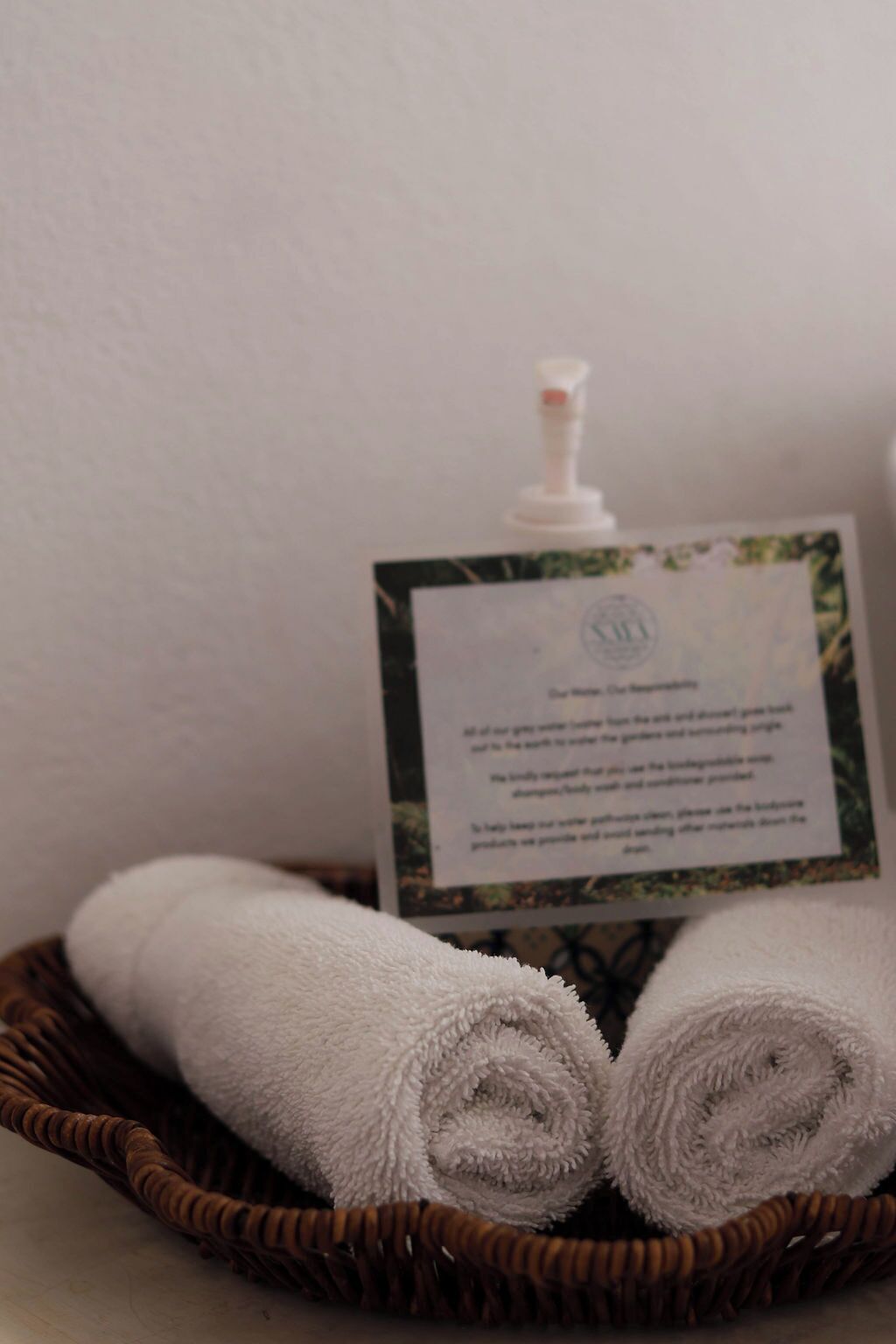 A white towel rolled in a wicker basket, a white bottle, and a card with text.