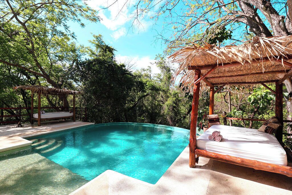 Ein Pool mit Holzterrasse, einer Holzkonstruktion als Sonnenschutz und einem Bett mit Matratze und Kissen.