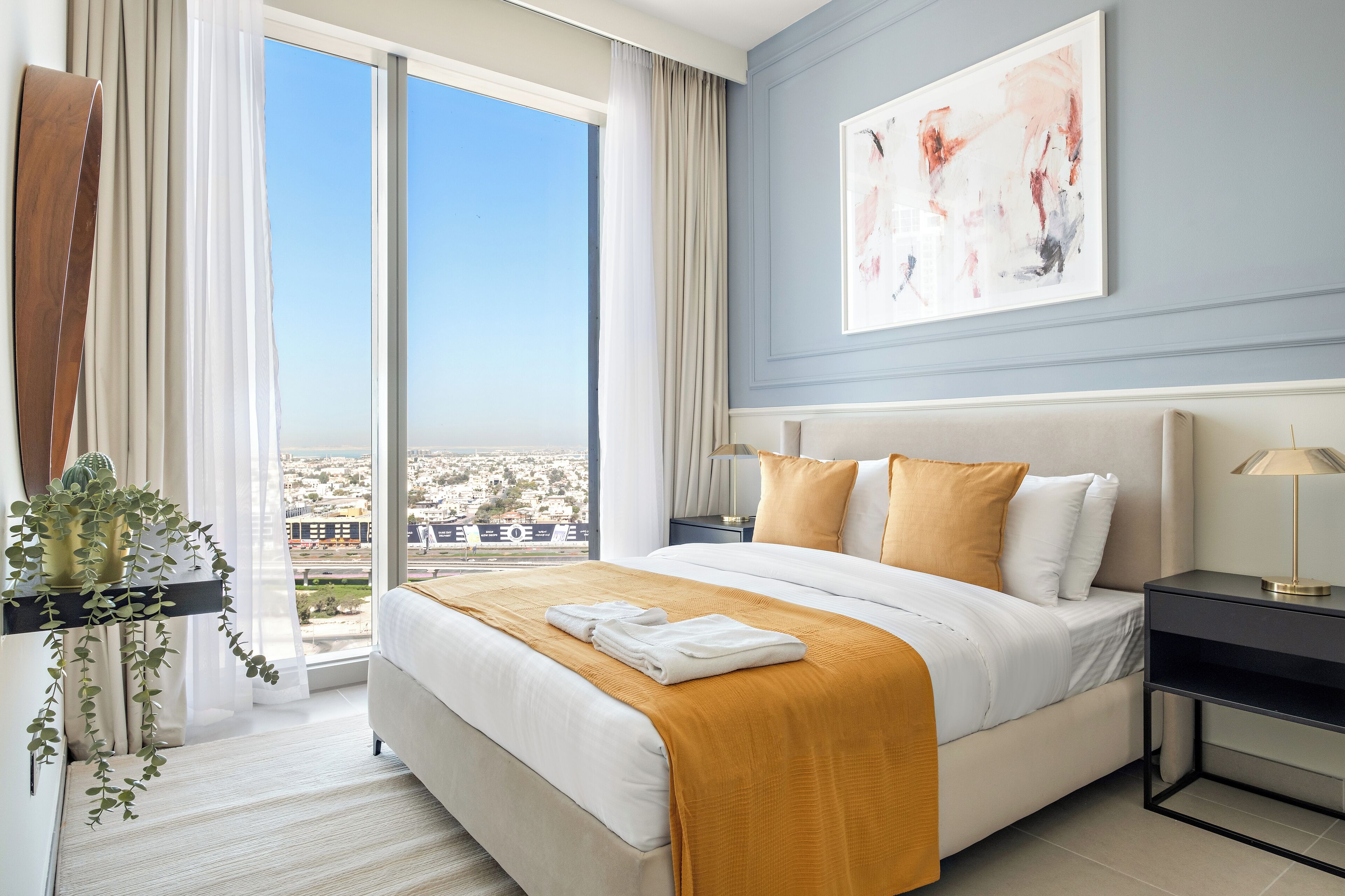 Una habitación de hotel moderna con una cama grande, una mesita de noche, una planta en maceta y vistas a la ciudad a través de amplias ventanas.